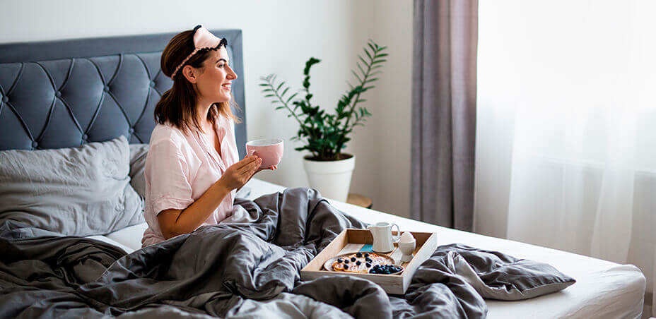 Brunette lady having coffee sitting in a bed with grey and white bedding and grey upholstered headboard